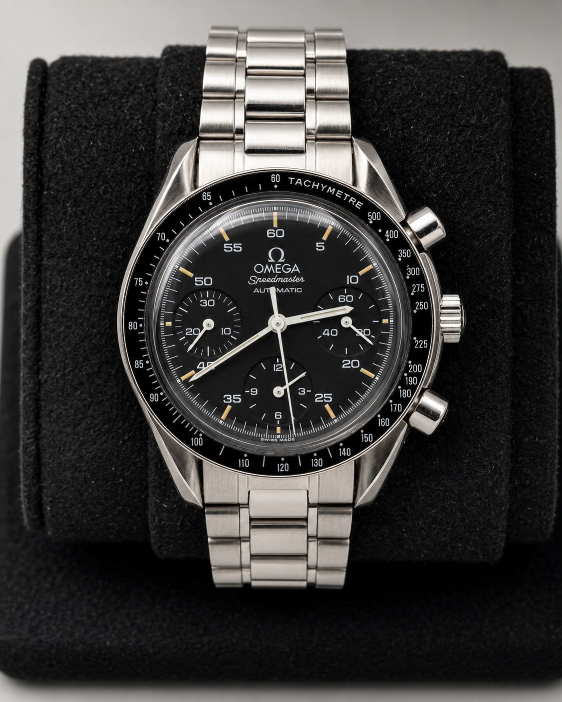 An Omega Speedmaster wristwatch with a black dial and black tachymeter bezel, featuring a silver stainless steel case and integrated bracelet, displayed on a black velvet watch cushion.