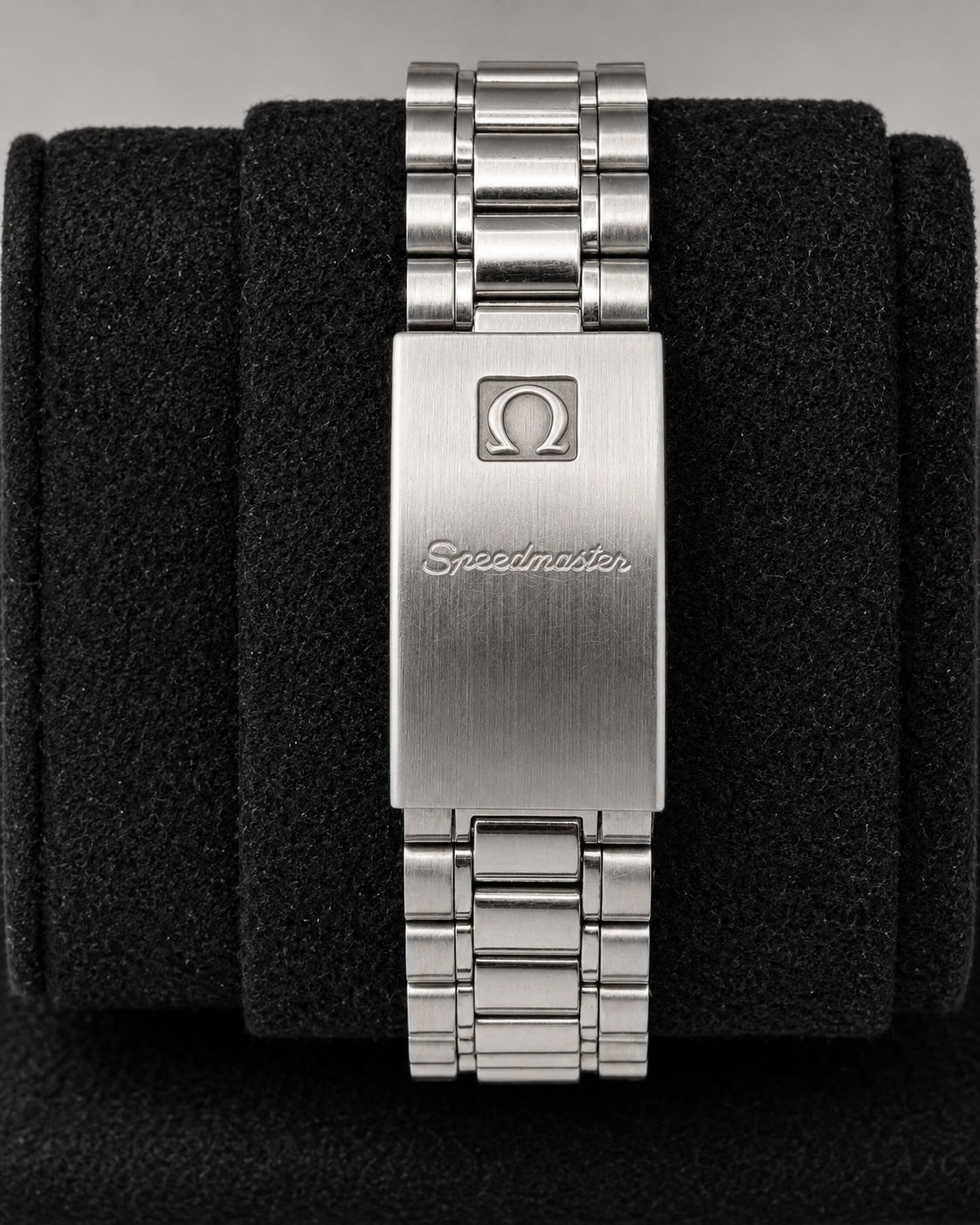 Silver stainless steel Omega Speedmaster watch band clasp with the Omega logo and 'Speedmaster' script, displayed on a black textured cushion.