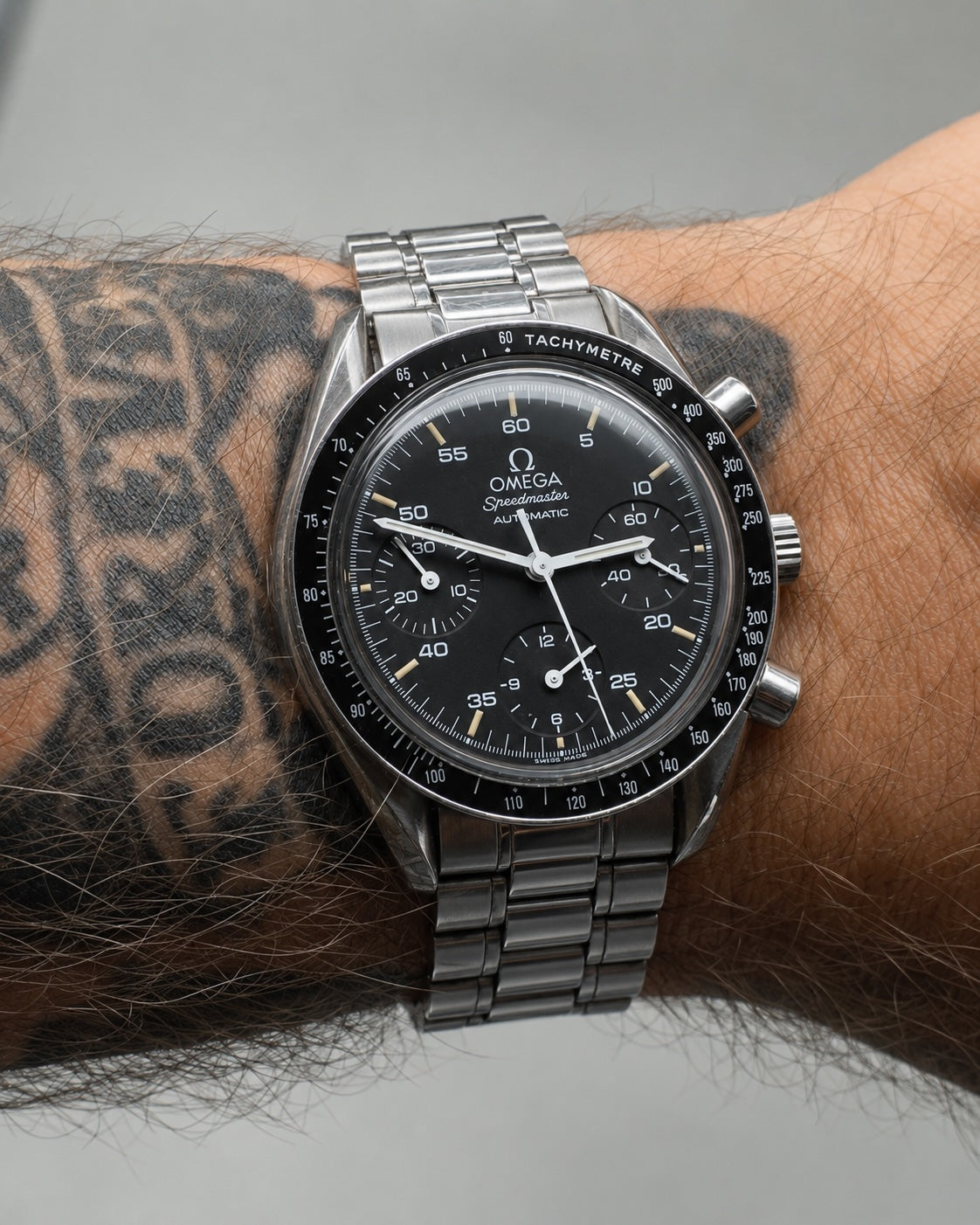 A close-up of an Omega x Cosmograph Daytona wristwatch with a black dial and silver stainless steel case and bracelet, worn on a person's wrist with a tattooed forearm visible.