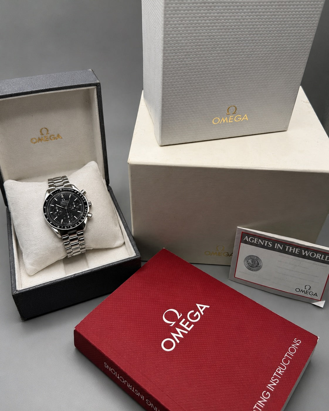 Omega wristwatch displayed in an open presentation box with a silver metal bracelet and black dial, alongside branded Omega packaging boxes, a red instruction booklet, and a certificate on a gray background.