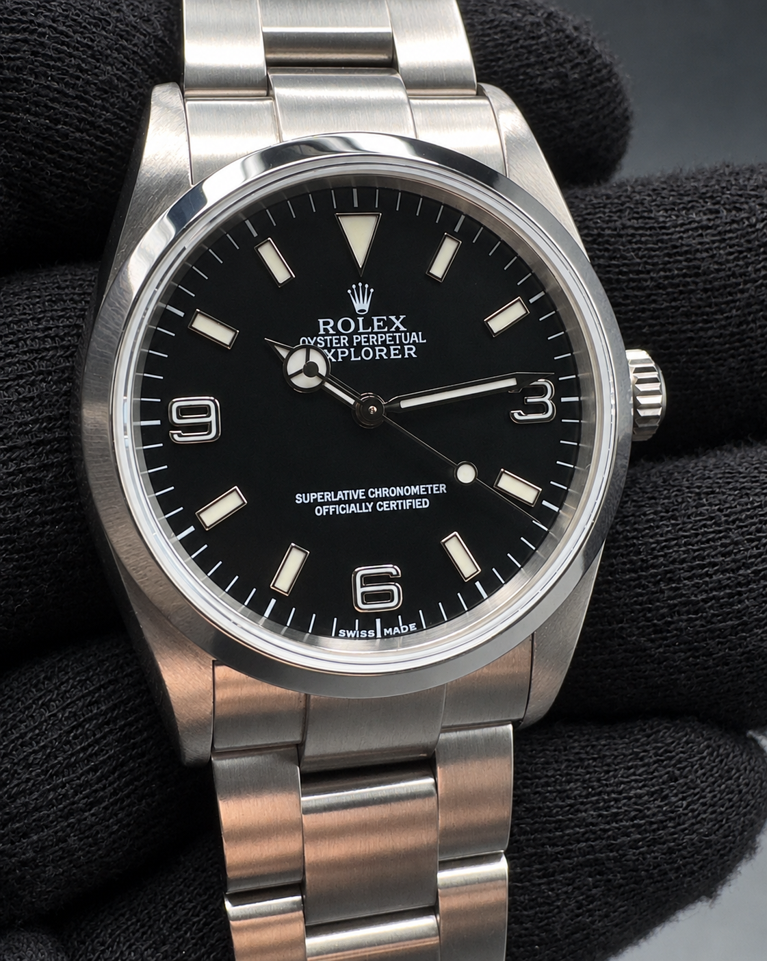 A Rolex Explorer wristwatch with a black dial and stainless steel Oyster bracelet, shown resting on a black textured surface.