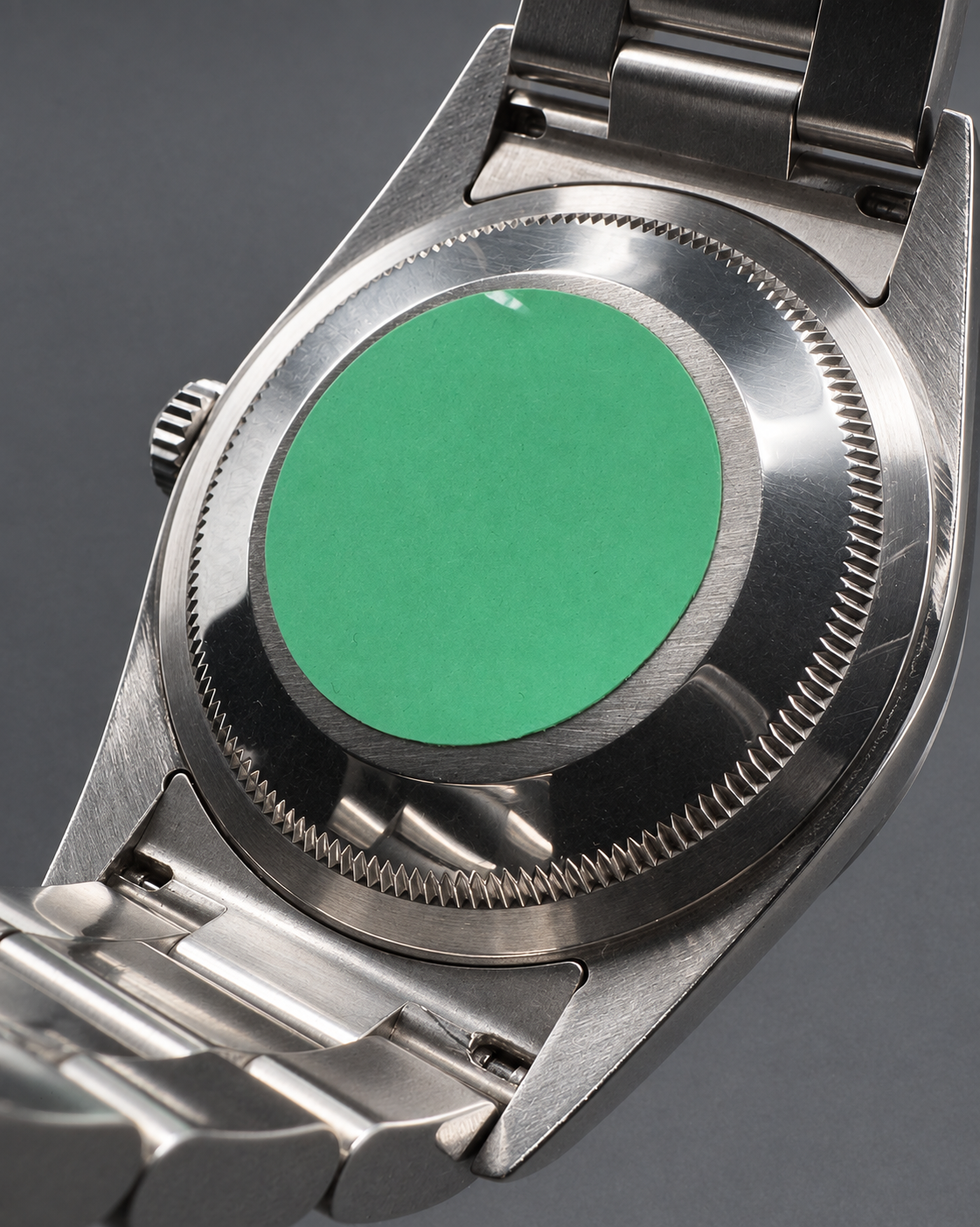 Close-up of the back of a stainless steel watch case with a metal bracelet, showing a brushed silver finish and a bright green circular insert or protective cover in the center.
