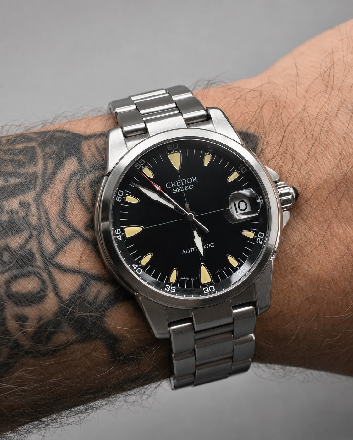 A close-up of a wrist wearing a Credor Geedor Auto Sync watch with a black dial, silver stainless steel case and bracelet, and yellow-gold indices.