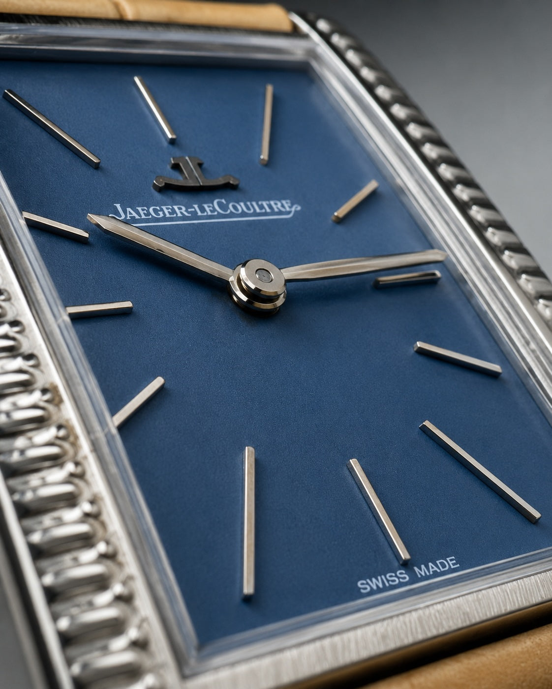 Jaeger-LeCoultre watch close-up showing a deep blue dial, silver-tone baton hour markers, and a silver-tone rectangular bezel with a textured outer edge.