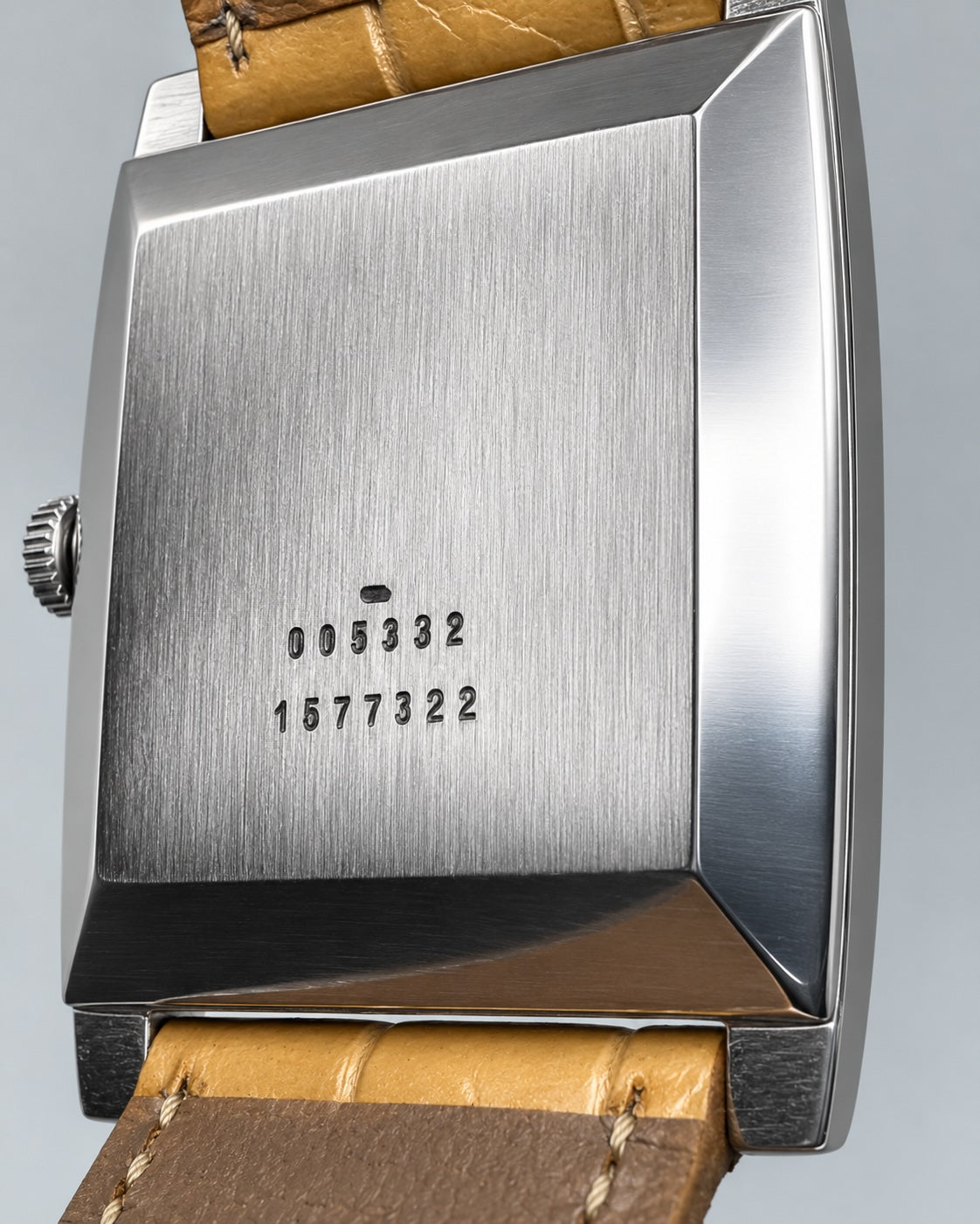 Close-up of the back of a rectangular stainless steel wristwatch case with a tan leather strap, showing engraved serial numbers on the brushed metal.