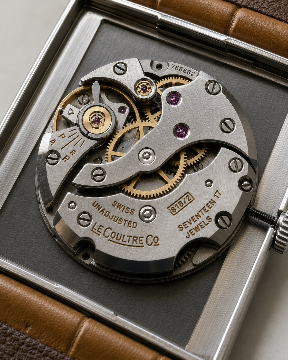 Close-up of a Le Coultre Co. Seventeen Jewels Swiss unadjusted mechanical watch movement, showing silver-toned gears, gold-toned components, and purple jewel bearings, set against a dark gray insert and brown leather strap.