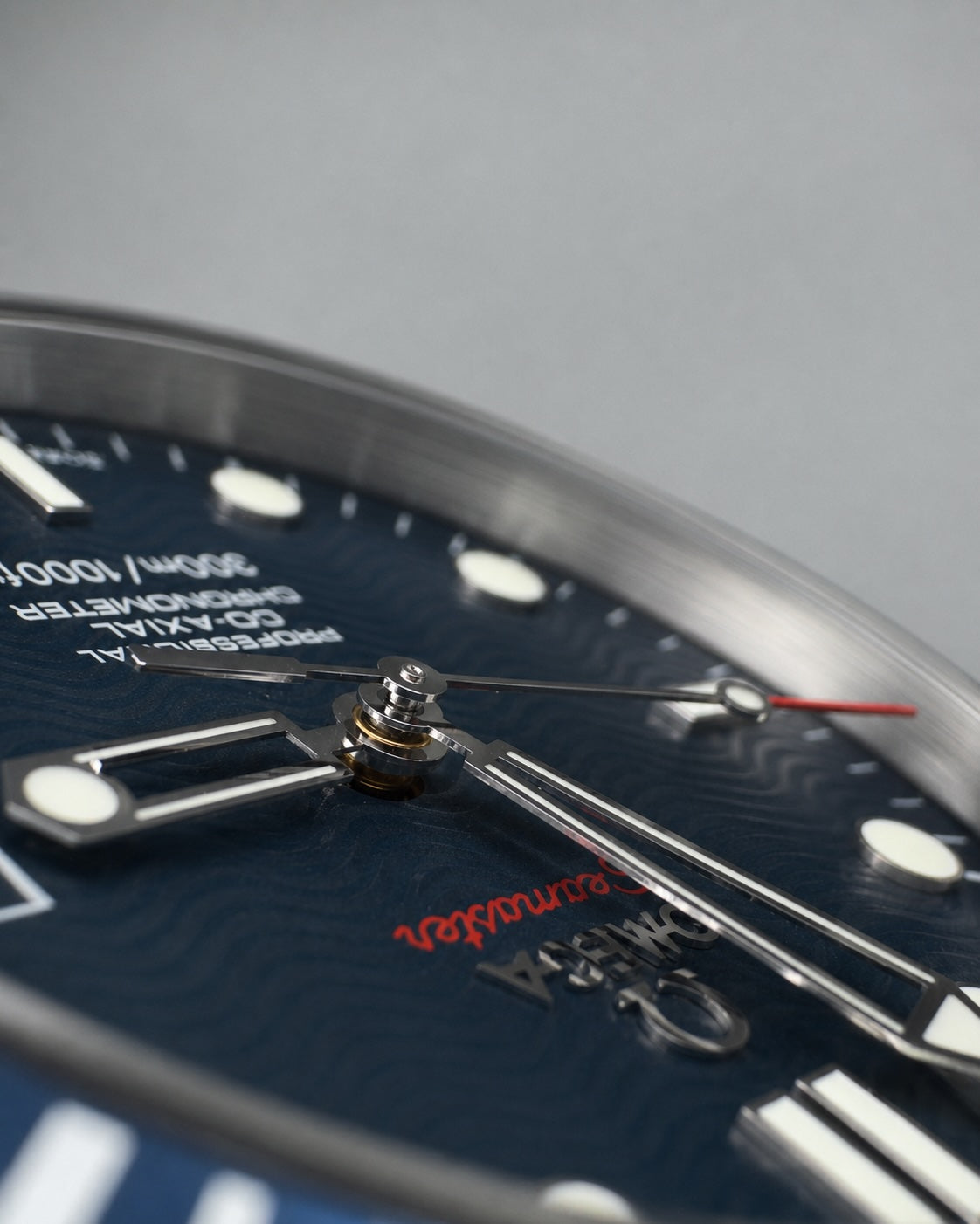 Close-up of a wristwatch dial with a dark blue textured face, silver-tone hands and markers, and a thin red seconds hand. The brand name 'Omega' is visible in red on the dial.