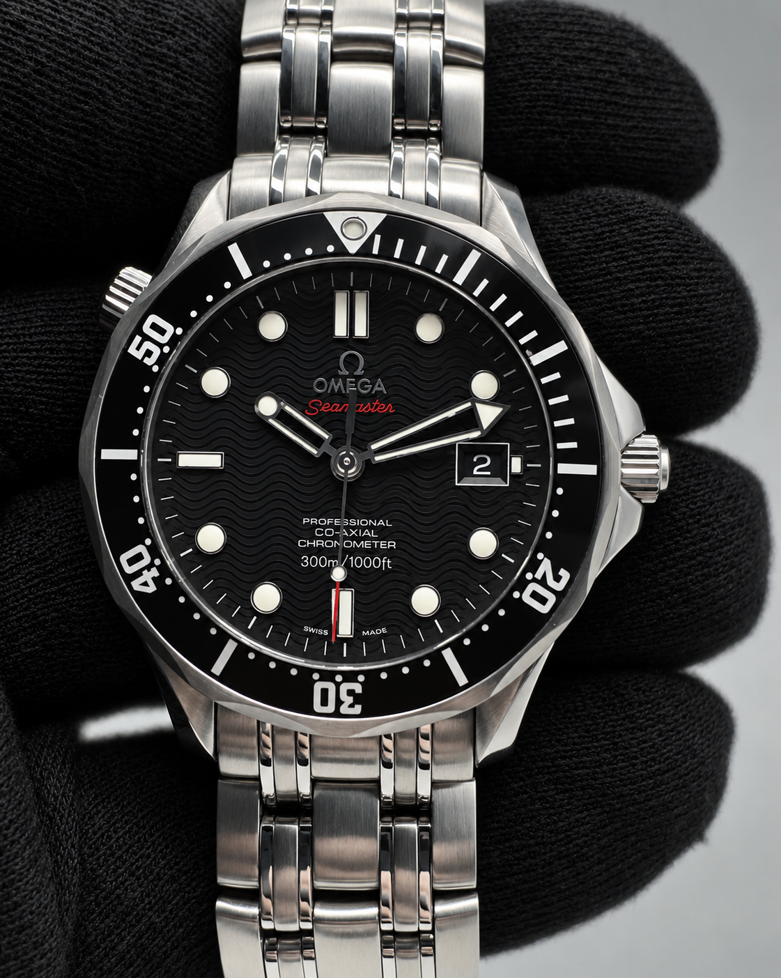 An Omega Seamaster wristwatch with a black dial and silver stainless steel bracelet, held by a black gloved hand against a neutral gray background.