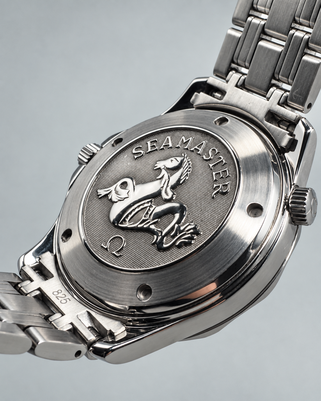 Close-up of the back of a stainless steel wristwatch showing the Sea-Master frog logo and brand name on the case, with part of the metal bracelet visible against a soft gray background.