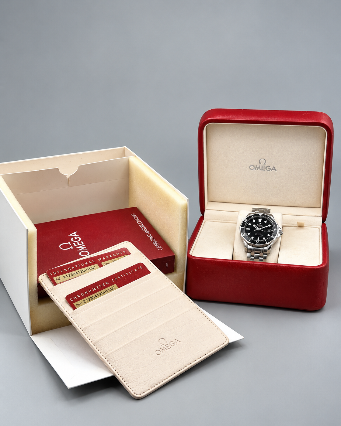 An Omega wristwatch with a black dial and silver metal case displayed in an open red presentation box, alongside an open cream-colored Omega box containing documentation and a matching cream card holder on a gray background.