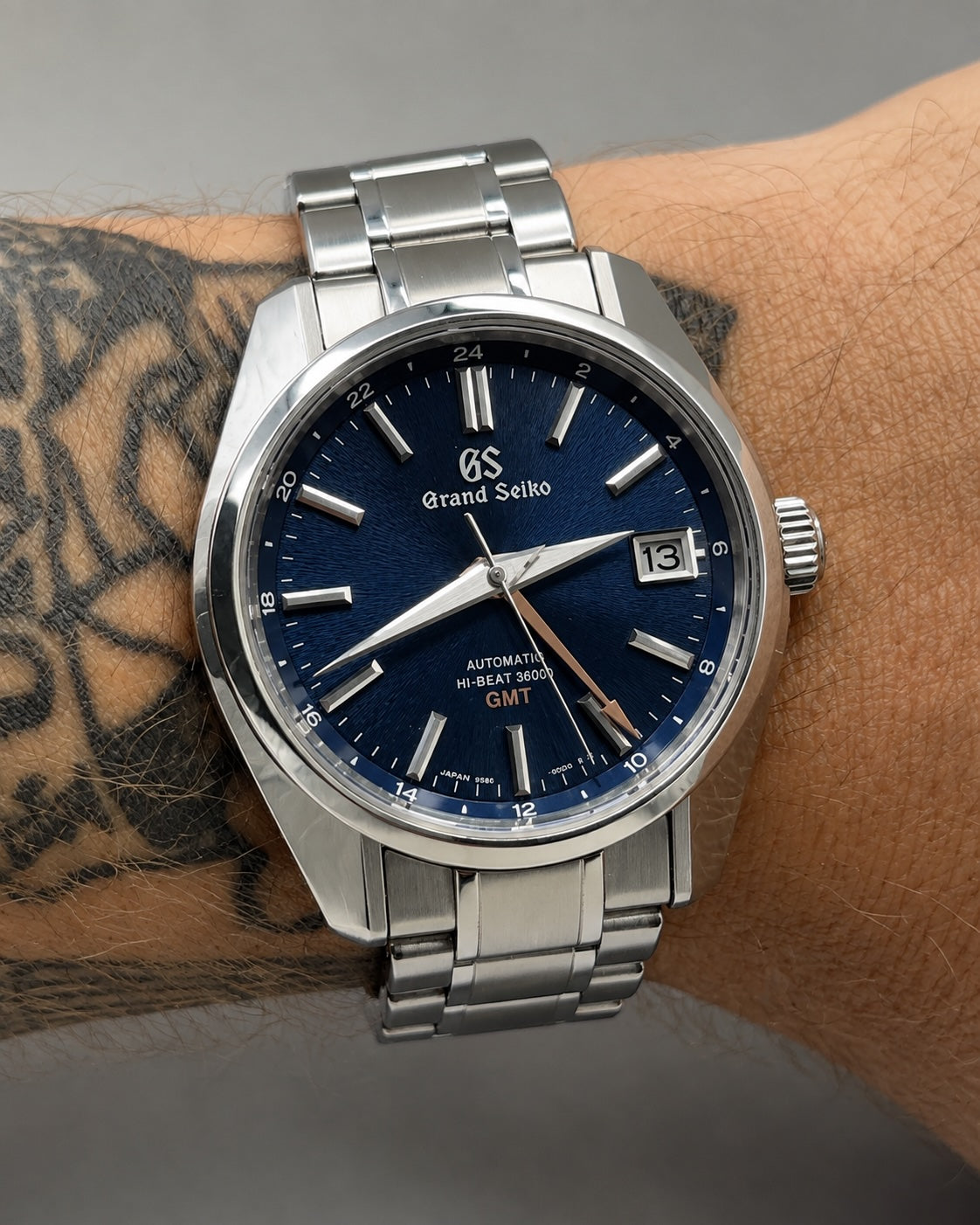 A close-up of a Grand Seiko wristwatch with a stainless steel link bracelet and a deep blue dial, worn on a person's tattooed wrist.