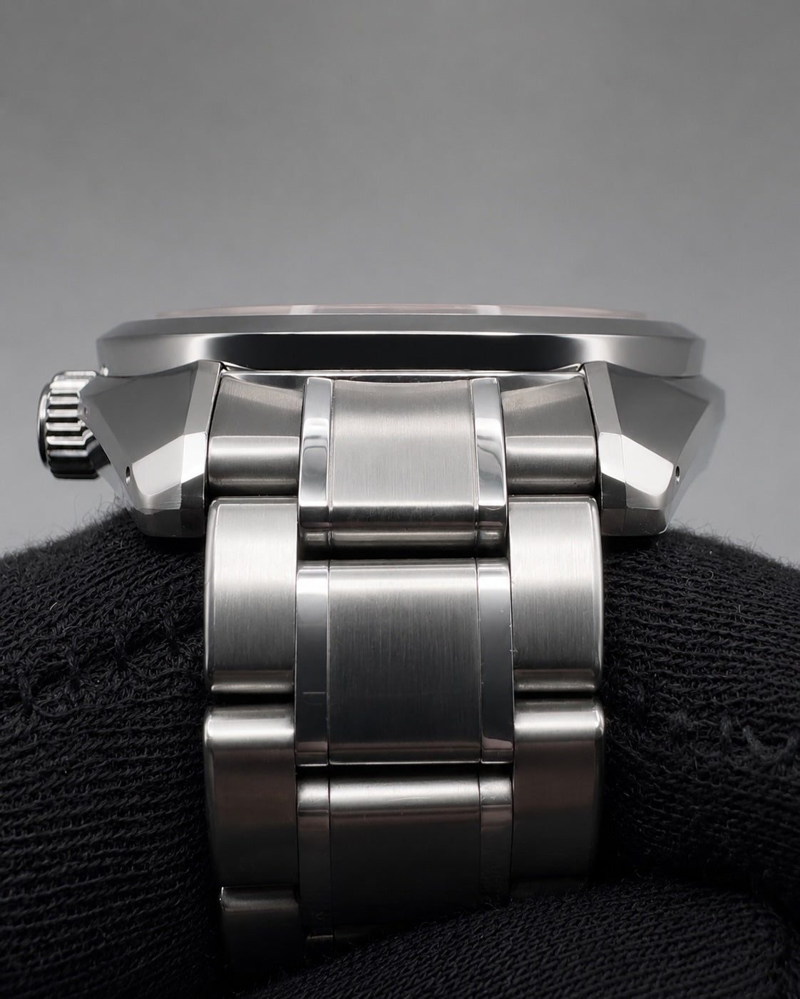 Close-up of the side and bracelet of a stainless steel watch resting on a black textured surface against a gray background.