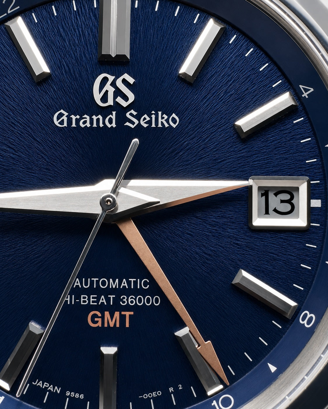 Close-up of a Grand Seiko automatic watch dial with a deep blue textured background, silver hour markers and hands, a date window showing 13, and orange GMT text.