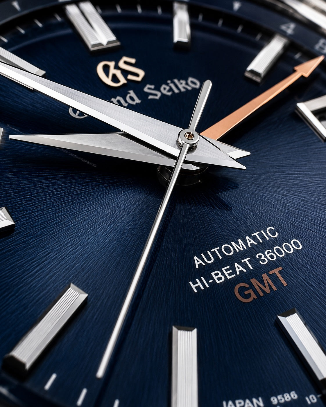 Close-up of a Grand Seiko automatic watch dial with a deep blue textured background, silver baton hour markers, silver hands, and a rose gold GMT indicator hand.