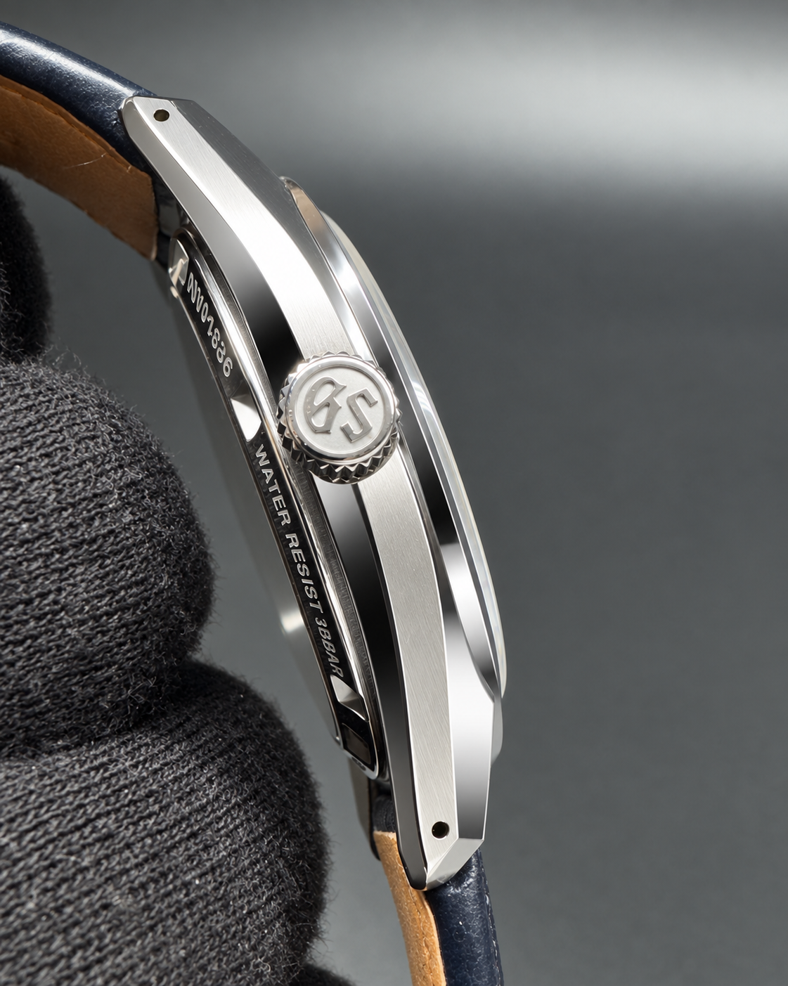 Close-up of a silver-toned watch case with a round crown accent and engraved details, attached to a dark blue leather strap with tan edges, held against a dark gray background.