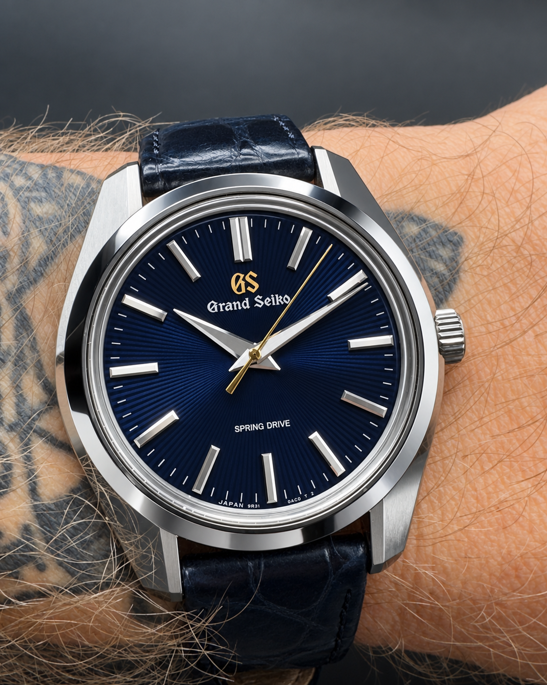 A close-up of a Grand Seiko wristwatch with a deep blue sunburst dial, silver stainless steel case, and a dark blue leather strap worn on a person's tattooed wrist.