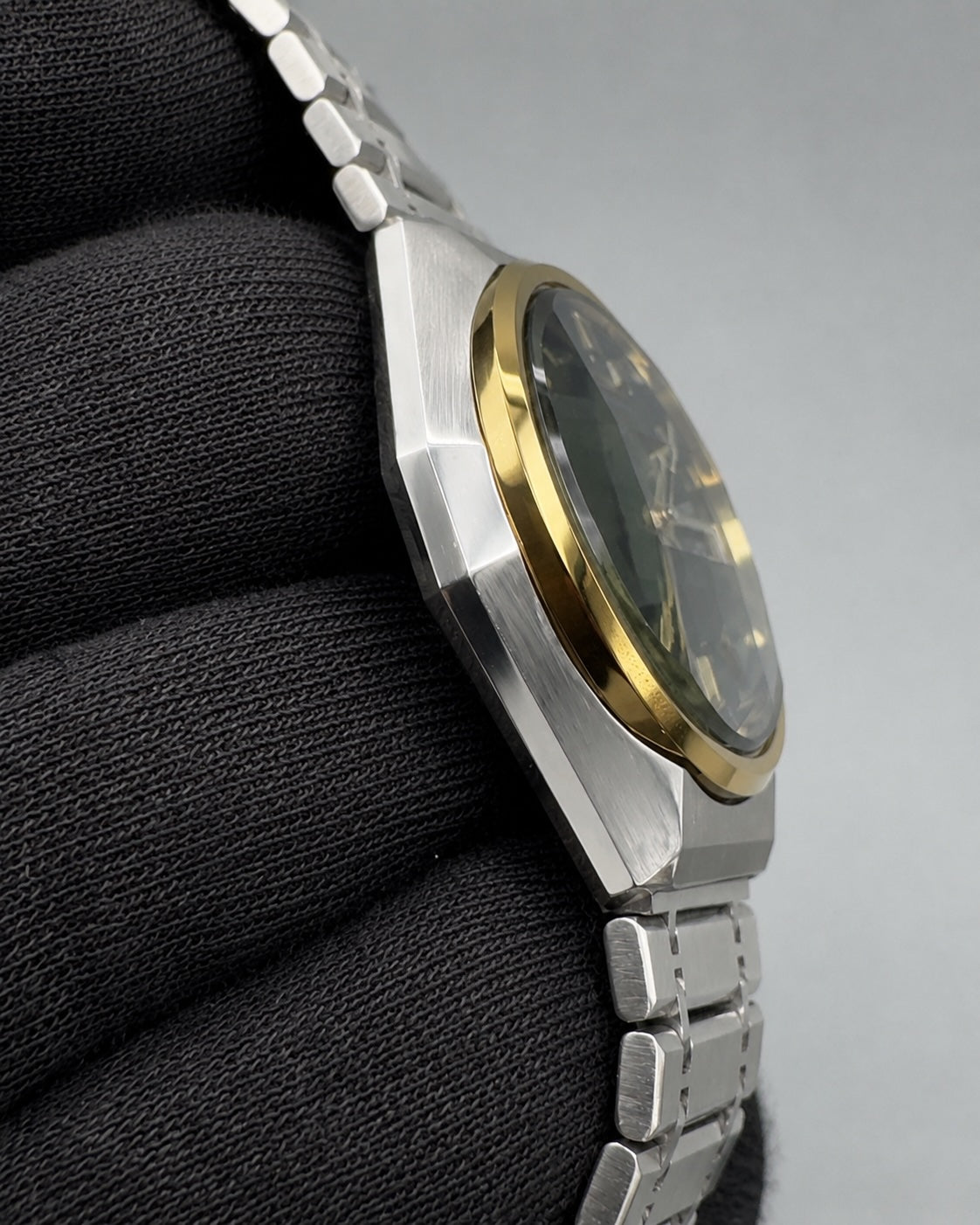 A close-up side view of a stainless steel watch with a gold bezel and dark dial, resting on a black textured surface against a gray background.