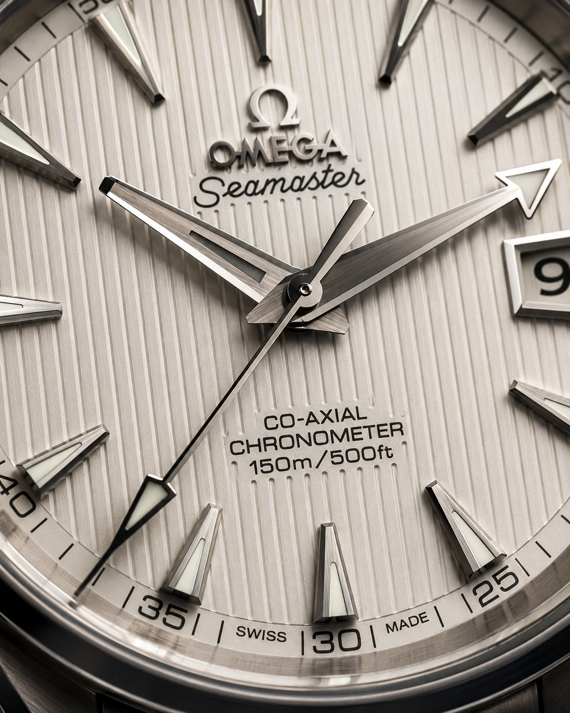 Close-up of an Omega Seamaster Co-Axial Chronometer watch dial, showing silver-toned hands, triangular hour markers, and the brand name on a light gray textured face.