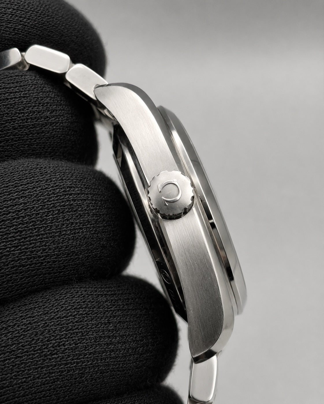 Close-up side view of a stainless steel wristwatch with a brushed metal case and link bracelet, resting on a black textured surface against a light gray background.