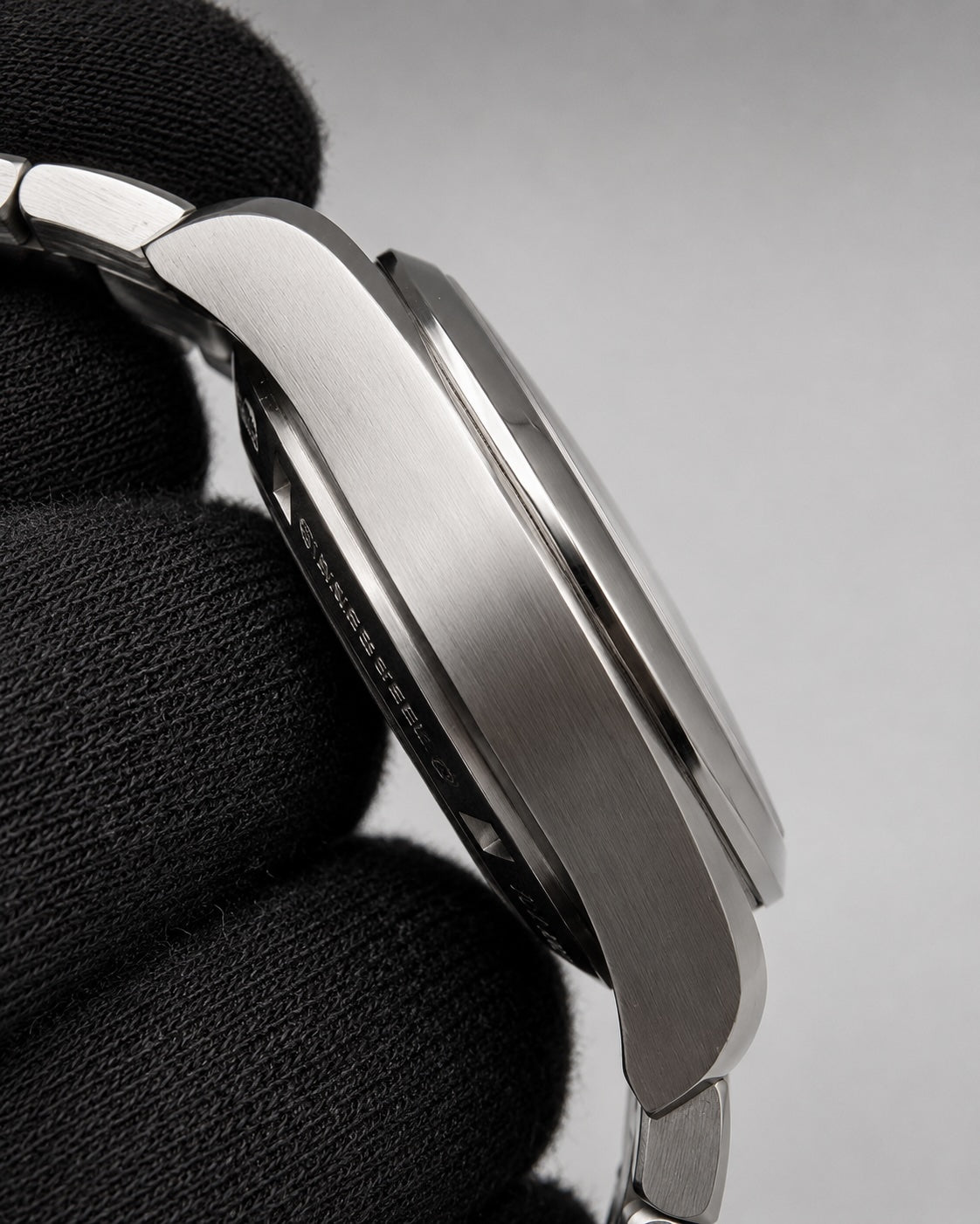 Close-up side view of a stainless steel watch case held against a black textured glove, showing brushed metal and polished edges on a light gray background.