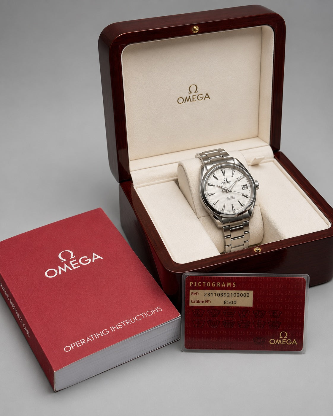 An Omega wristwatch displayed inside an open dark wood presentation box, with the red Omega box and an instruction booklet placed beside it on a light gray background.