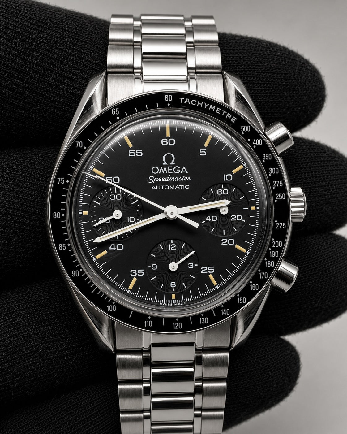 Omega Speedmaster Automatic wristwatch with a black dial, black tachymetre bezel, and silver stainless steel case and bracelet, shown resting on a black textured surface.