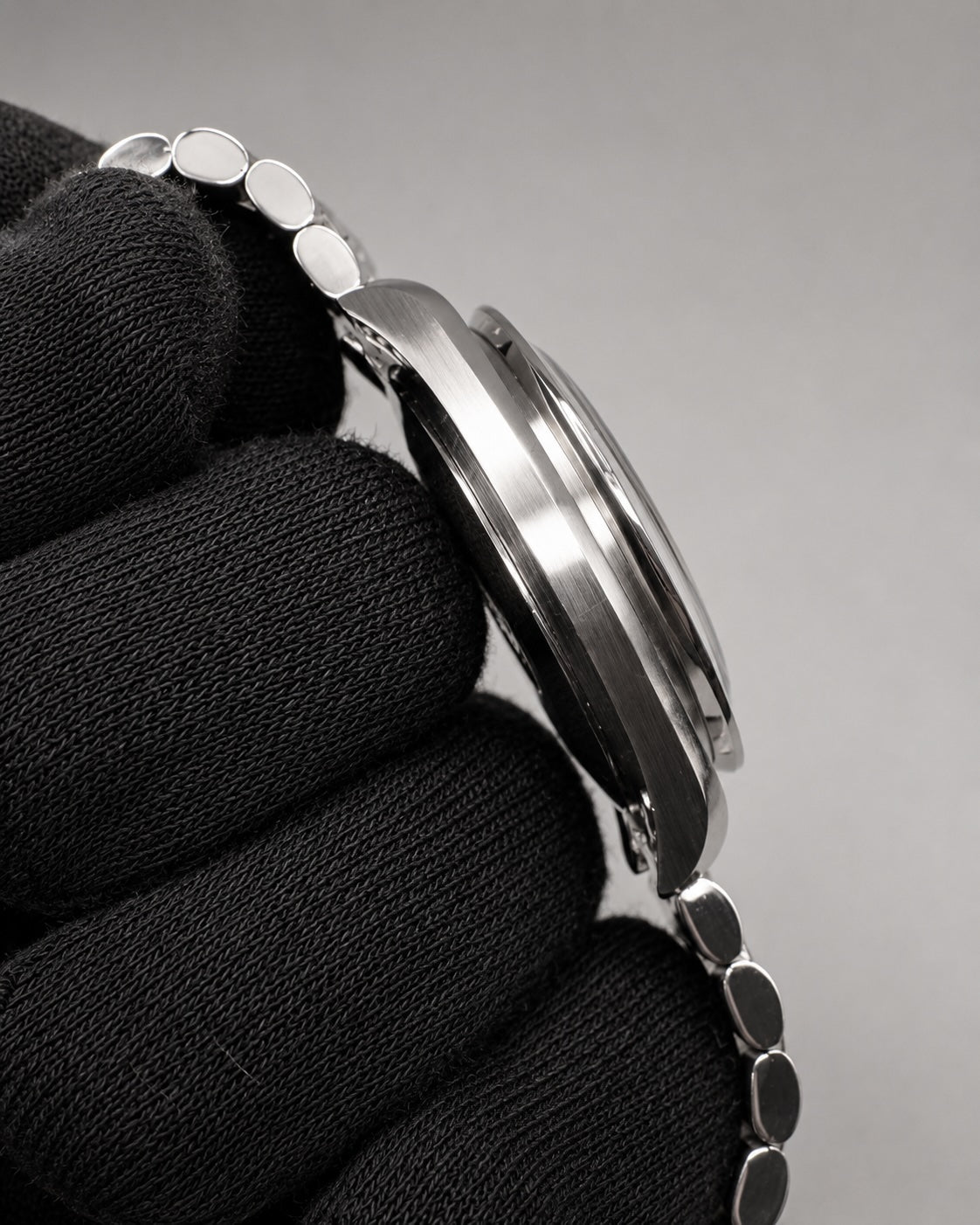 A close-up side view of a silver-toned metal watch with a matching link bracelet, held against a black textured fabric background on a neutral gray studio backdrop.