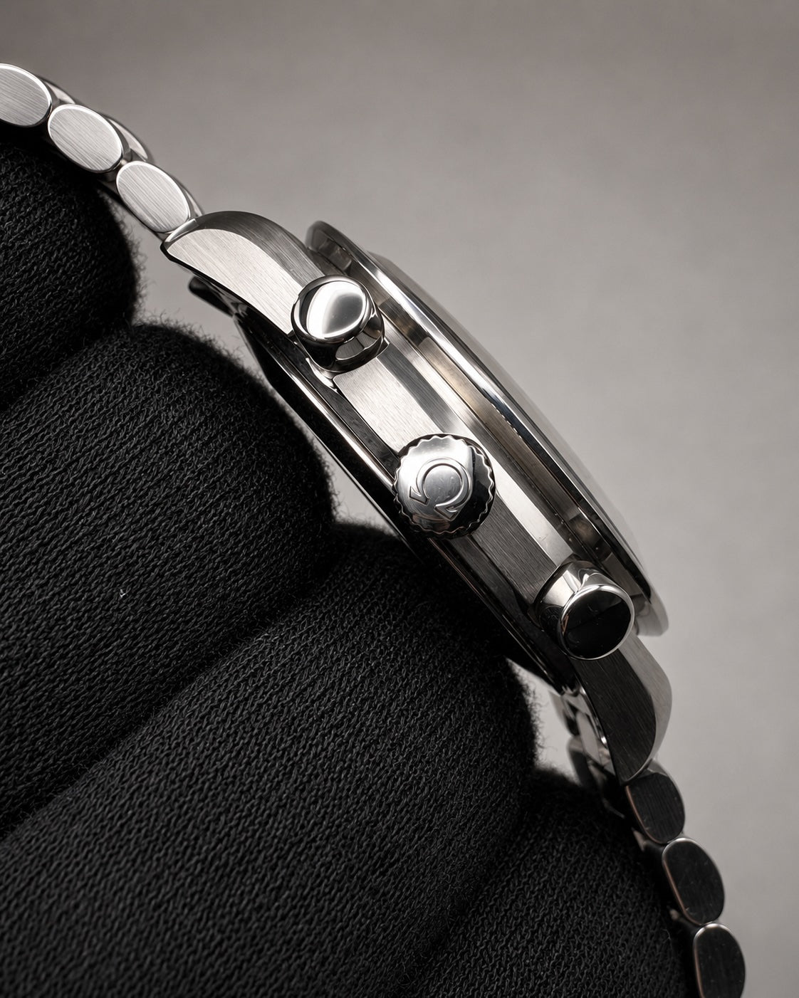Close-up side profile of a stainless steel wristwatch with a metal link bracelet, resting on a textured black fabric surface against a neutral gray background.