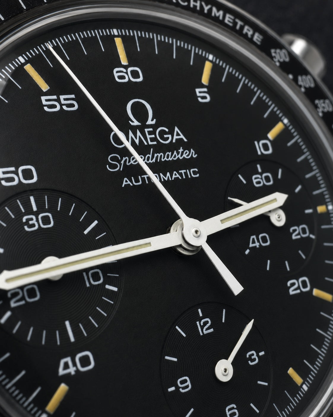 Close-up of an Omega Speedmaster Automatic watch dial, showing a black textured bezel, white hands and subdials, and yellow index markers.