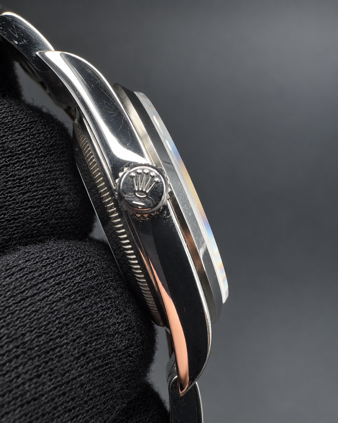 Close-up side view of a luxury wristwatch with a polished silver-tone case, a crown jewel featuring a crown motif, and a black textured strap on a dark gray background.