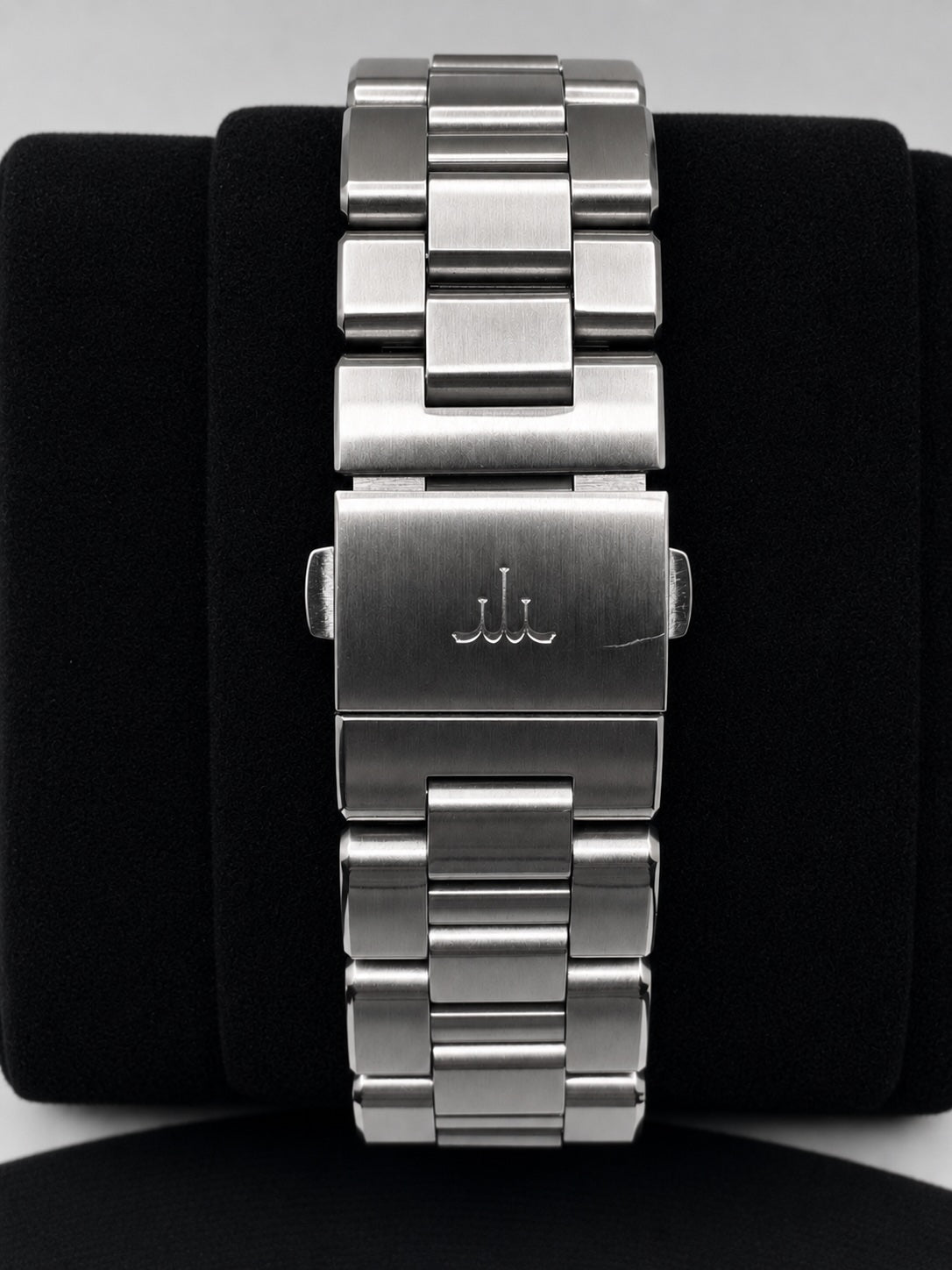 A close-up of a silver stainless steel link watch band with a brushed metal clasp engraved with a stylized 'JJ' logo, displayed on a black velvet watch stand.
