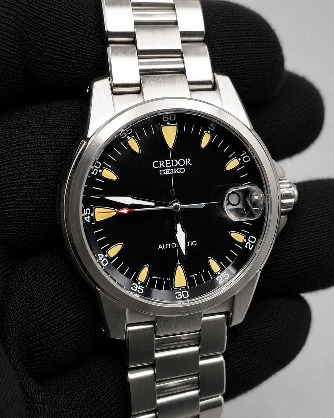 A stainless steel Credor Seiko wristwatch with a black dial and yellow luminescent markers, held by a hand wearing a black glove.