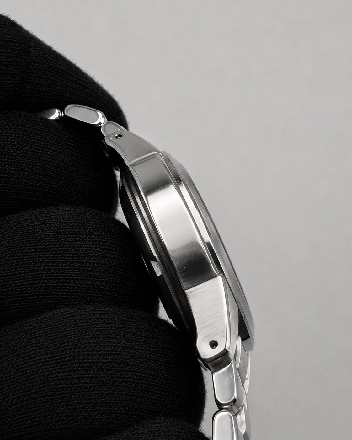Close-up side profile of a polished silver-tone metal watch bracelet held against a black textured fabric background, on a light gray studio background.