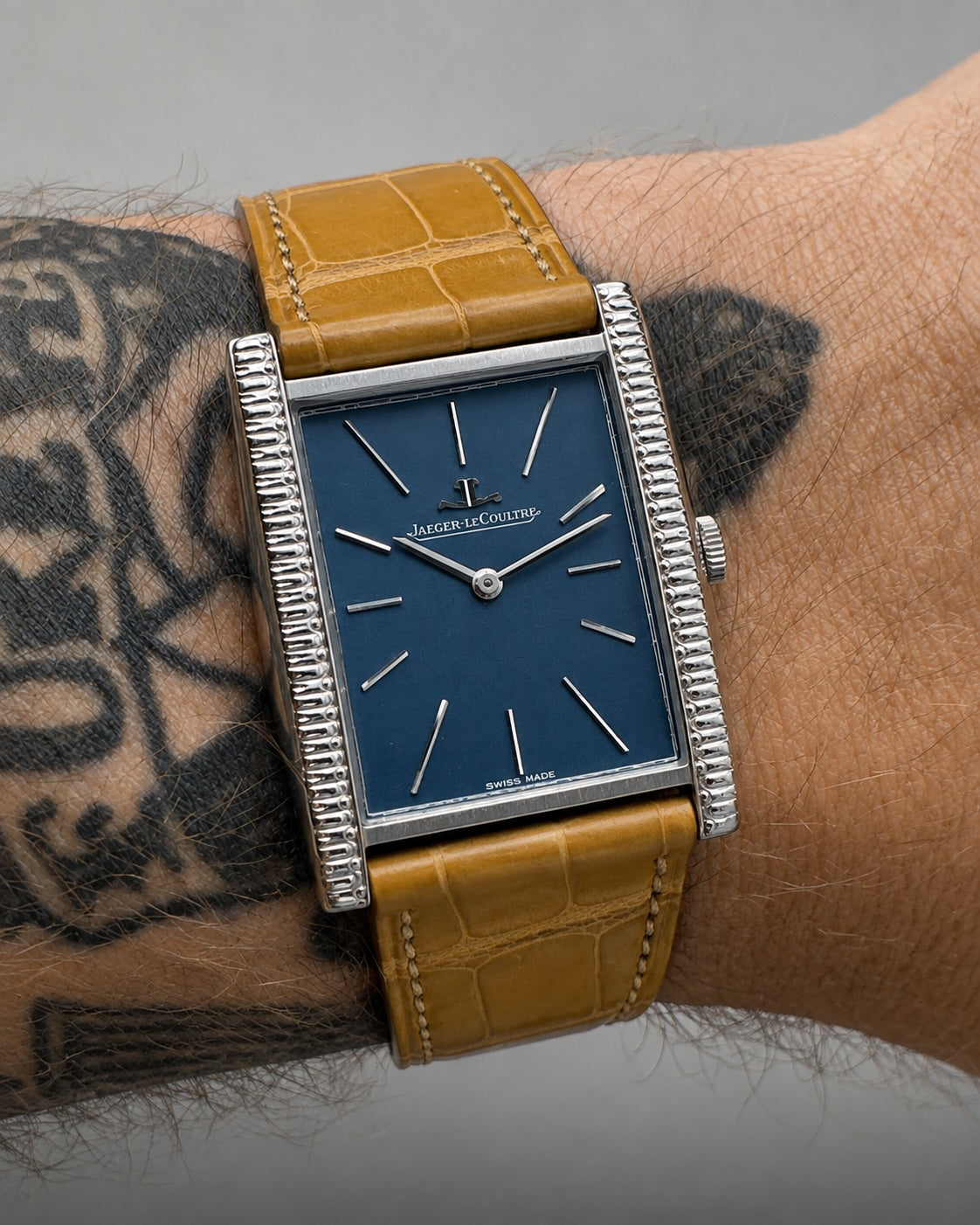 A close-up of a vintage-style wristwatch with a rectangular blue dial, silver-tone bezel, and tan leather strap, worn on a tattooed wrist against a neutral gray background.