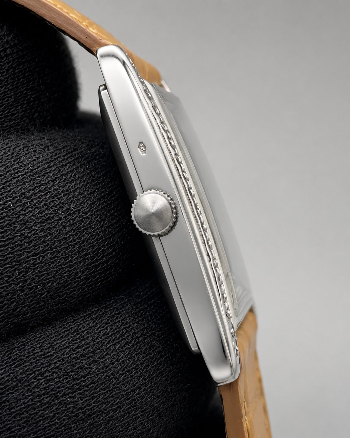Close-up side view of a silver-toned watch with a tan leather strap, resting on a black textured surface against a light gray background.