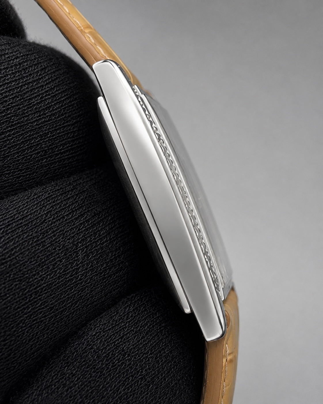 Close-up side view of a silver-toned watch case with a tan leather strap, resting on a black textured surface against a gray background.