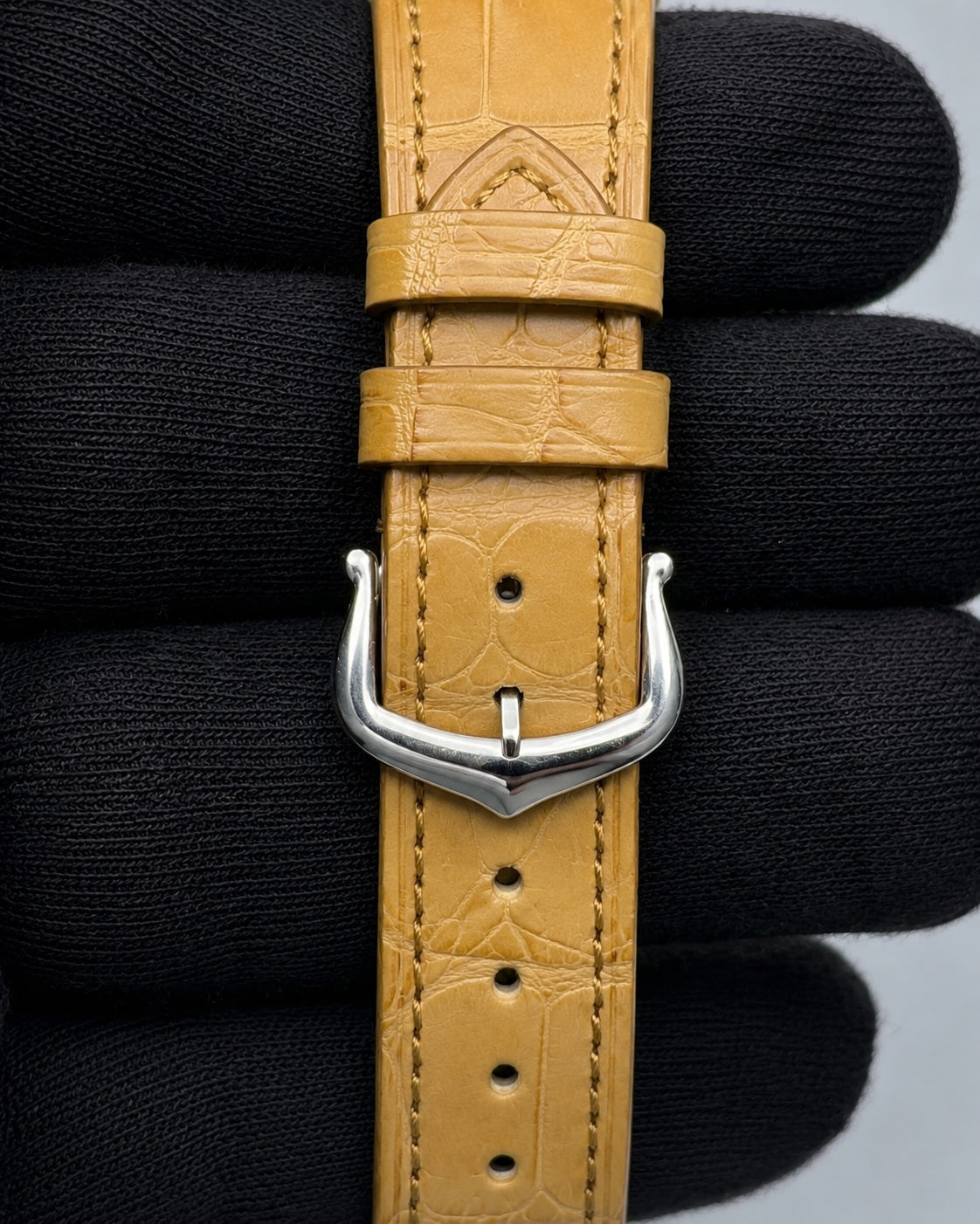 A close-up of a tan leather watch strap with an alligator or crocodile-embossed texture and a polished silver-tone buckle, held against a black knitted background.