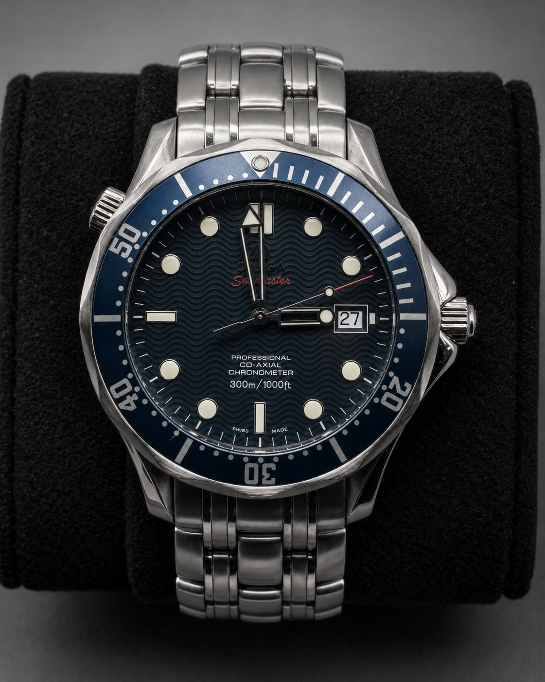 A stainless steel wristwatch with a blue dial and blue bezel, displayed on a black velvet watch cushion against a dark gray background.