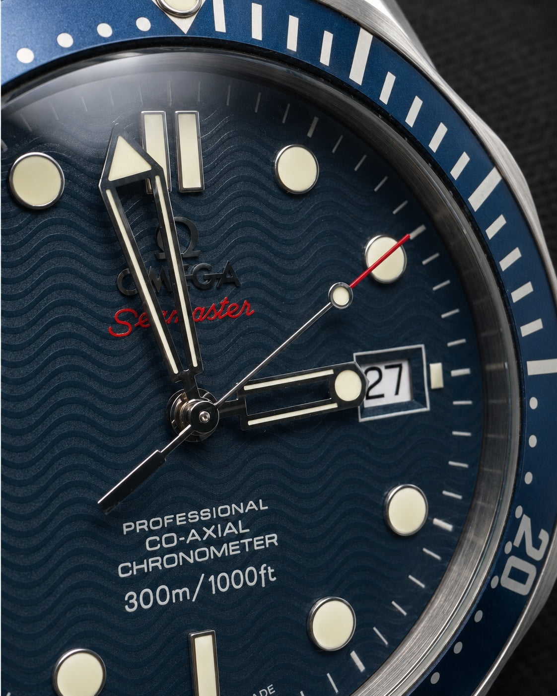 Close-up of an Omega Seamaster watch dial and bezel, showing a dark blue wavy textured dial with silver-tone hands, luminous hour markers, a date window, and a blue rotating bezel.