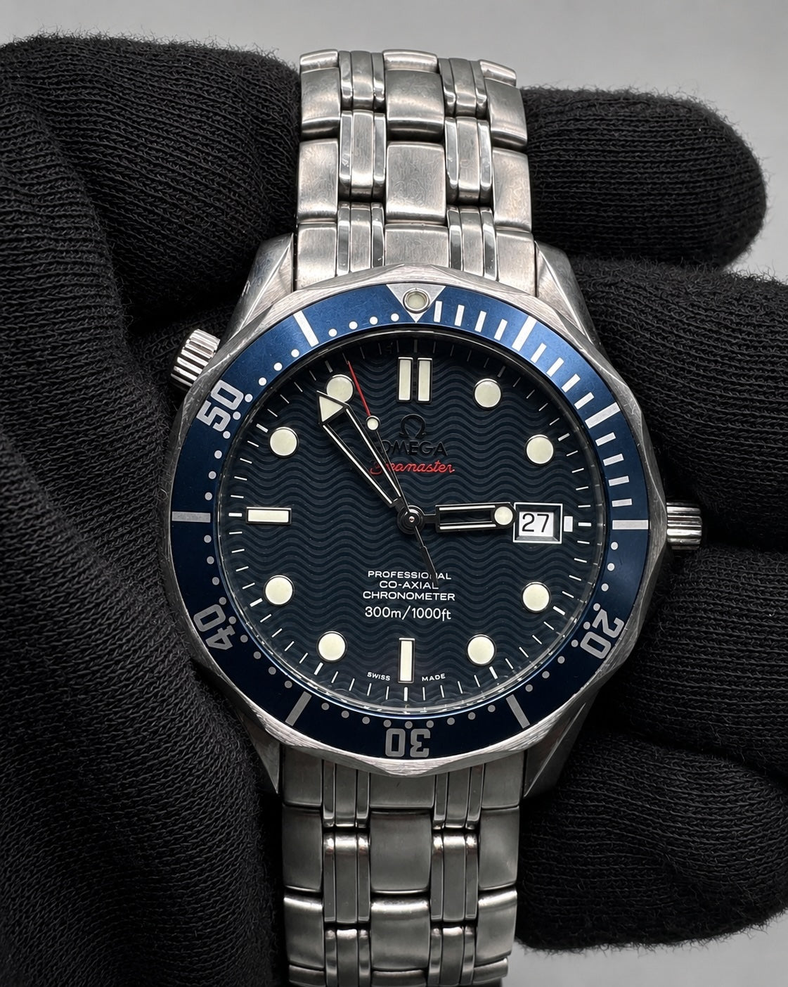 A stainless steel wristwatch with a blue dial and blue bezel, held in a black textured glove. The watch features a blue wave-pattern dial, silver-tone stainless steel case and link bracelet, and a blue bezel with white markings.