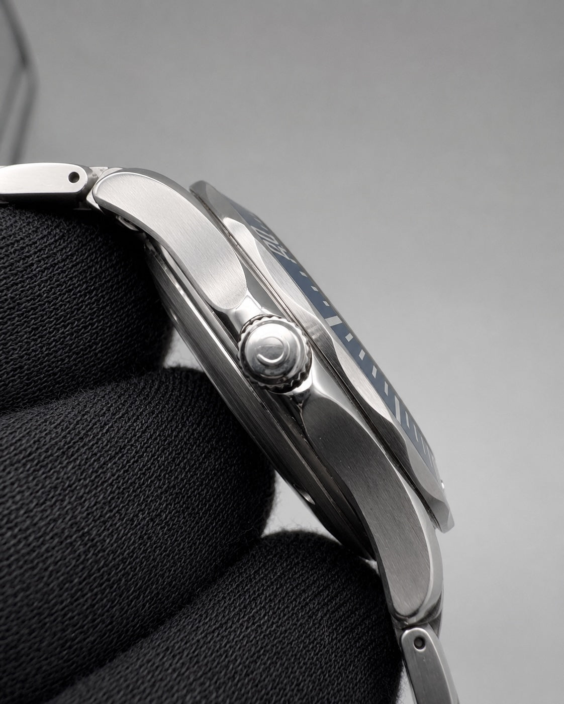 Close-up side profile of a stainless steel wristwatch held against a black textured fabric, showing the case, crown, and part of the blue dial on a gray background.
