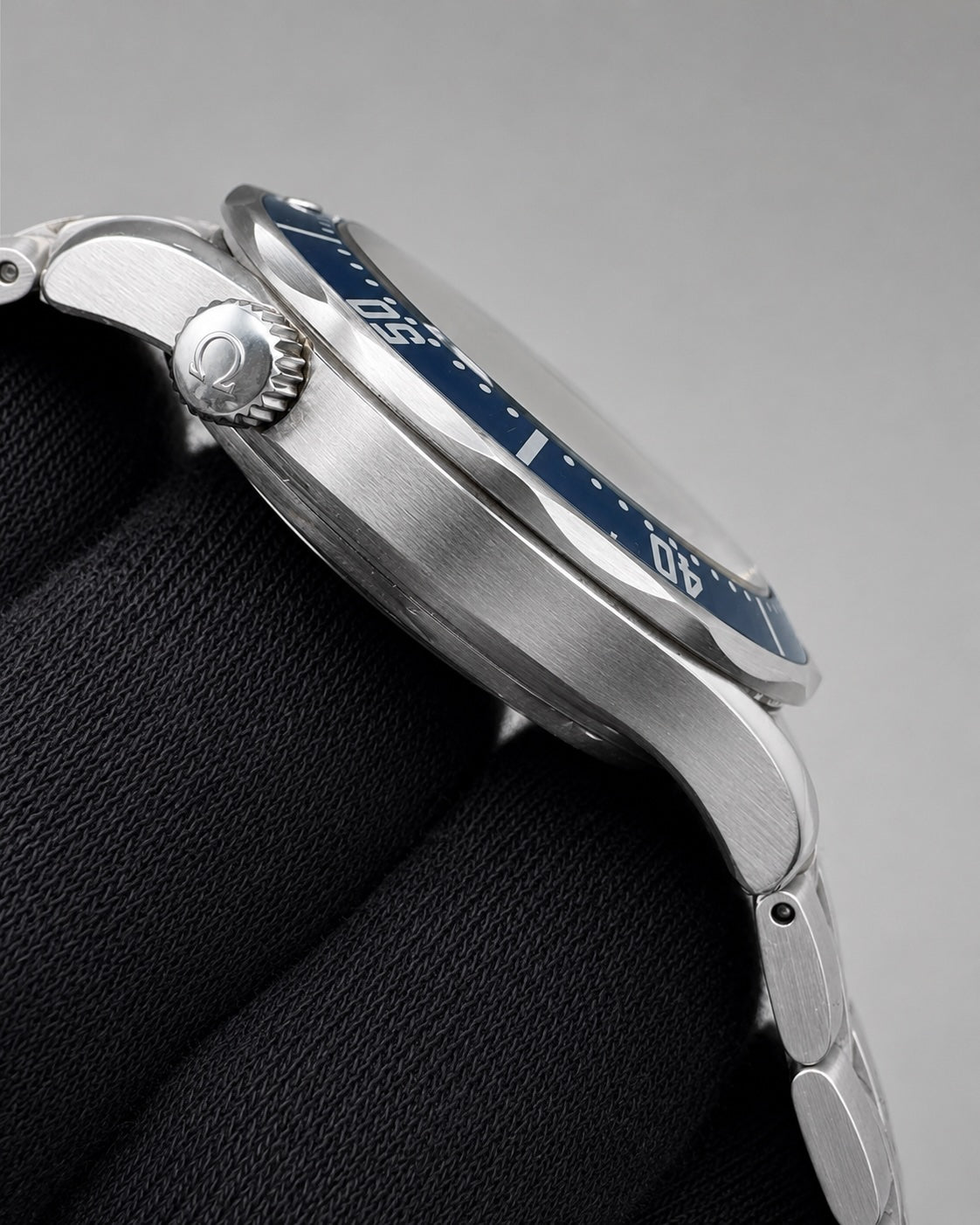 Close-up side view of a stainless steel wristwatch with a blue dial bezel and silver metal bracelet, resting on a black textured surface against a gray background.
