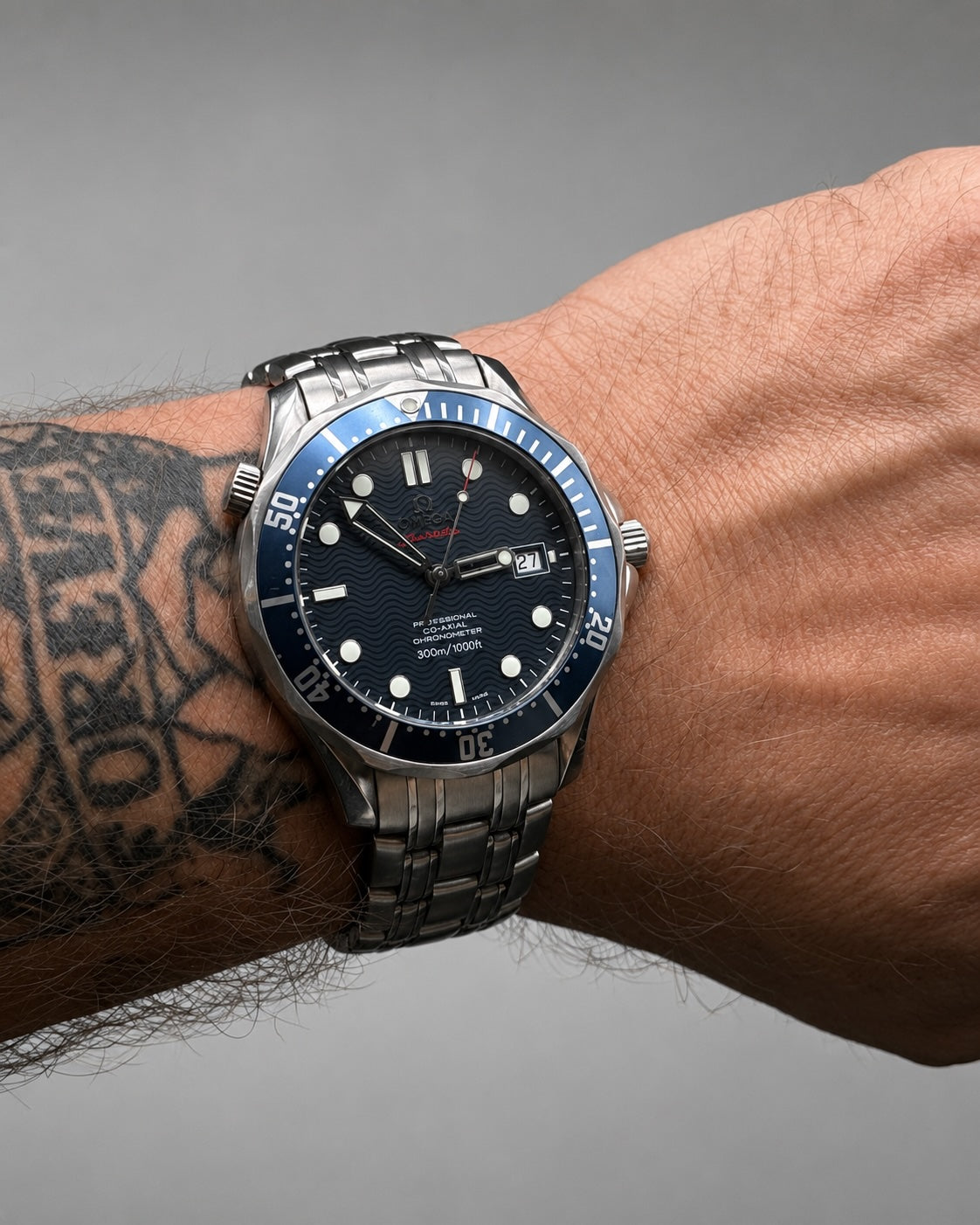 A stainless steel wristwatch with a dark blue dial and blue bezel, worn on a person's tattooed wrist against a gray background.