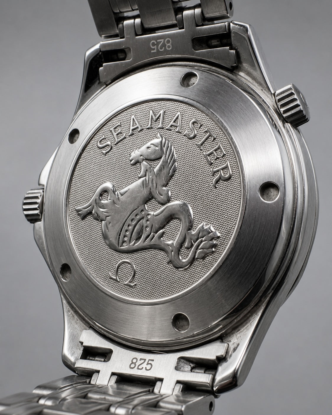 Close-up of the back of a stainless steel wristwatch showing the Seamaster case with the brand's winged anchor logo and 'SEAMASTER' text, attached to a metal link bracelet on a gray background.