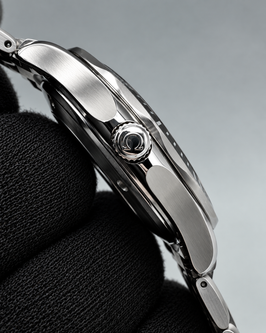 Close-up side profile of a stainless steel wristwatch held against a black textured fabric, showing the polished case, crown, and part of the metal bracelet on a soft gray background.