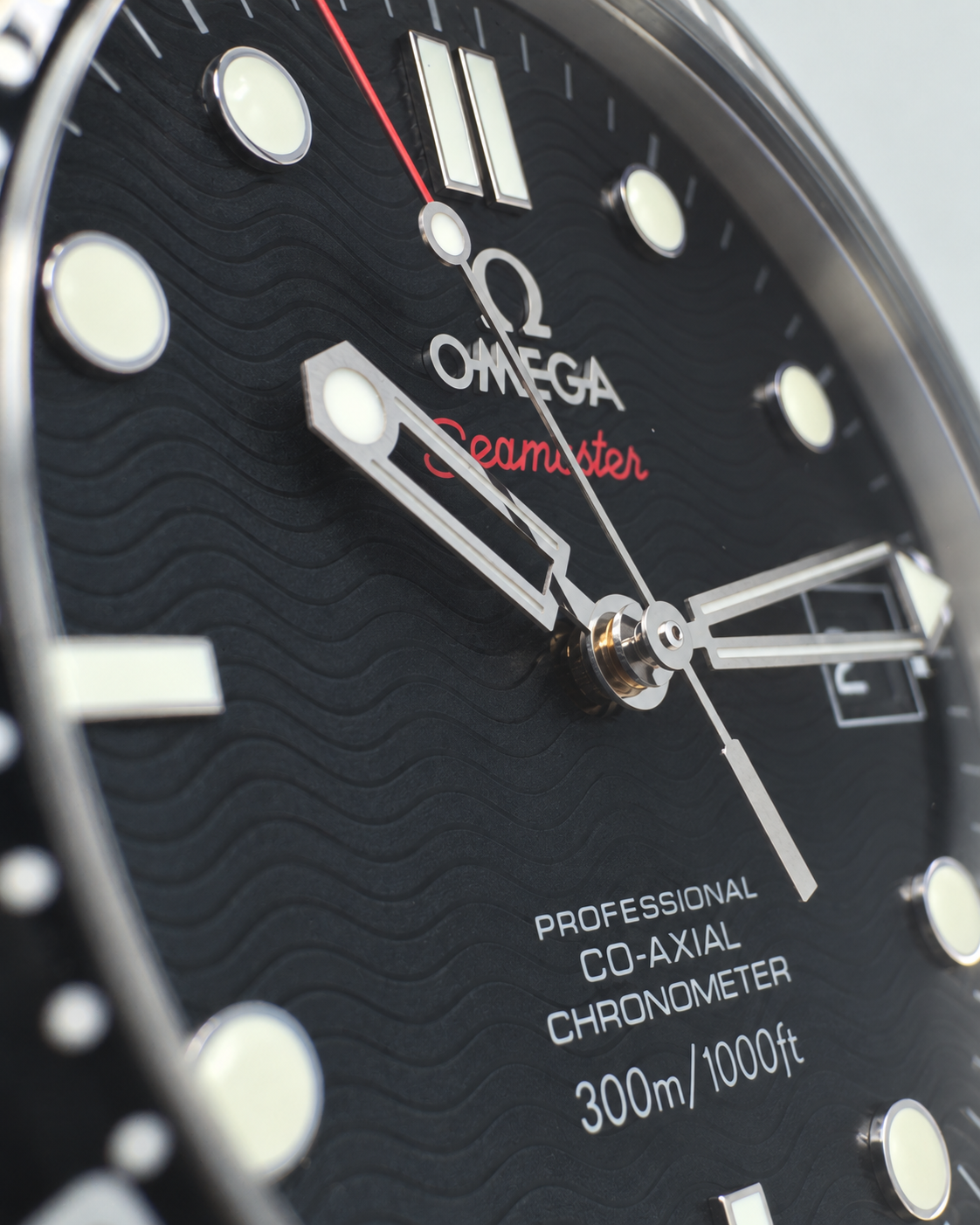 Close-up of an Omega Seamaster wristwatch dial, showing a black textured wave-patterned face with silver-tone hands, luminous hour markers, a red Seamaster script logo, and white text indicating 300m/1000ft.