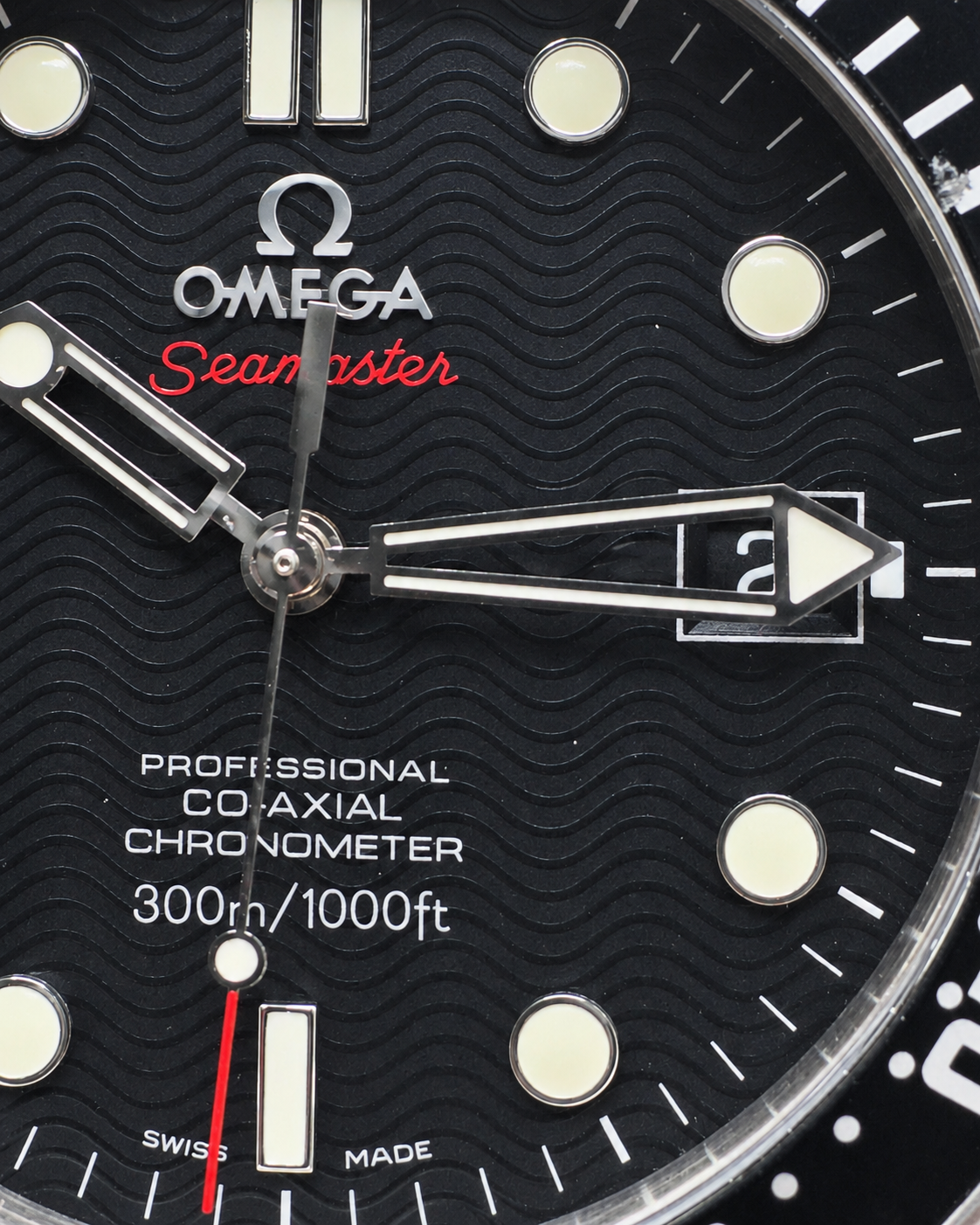 Close-up of an Omega Seamaster watch dial with a black textured wave-patterned bezel and black dial, featuring silver-tone hands, luminous hour markers, and red 'Seamaster' script.