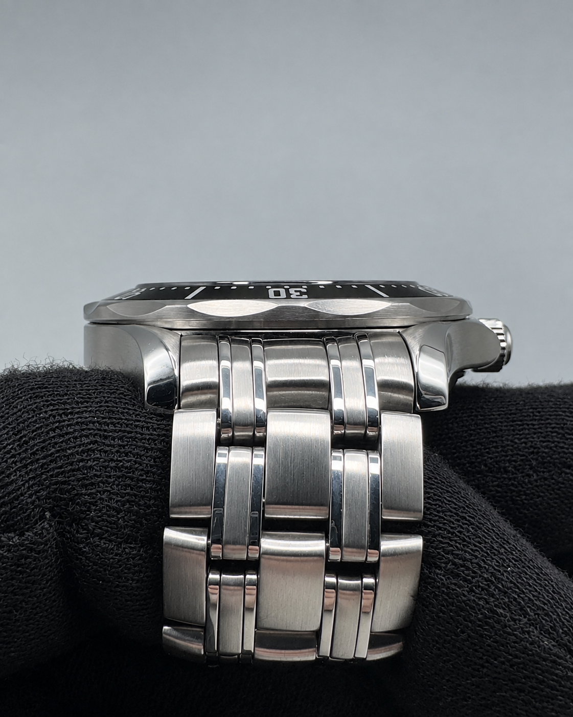A close-up of a stainless steel watch bracelet with brushed and polished links, resting on a black textured surface against a neutral gray background.