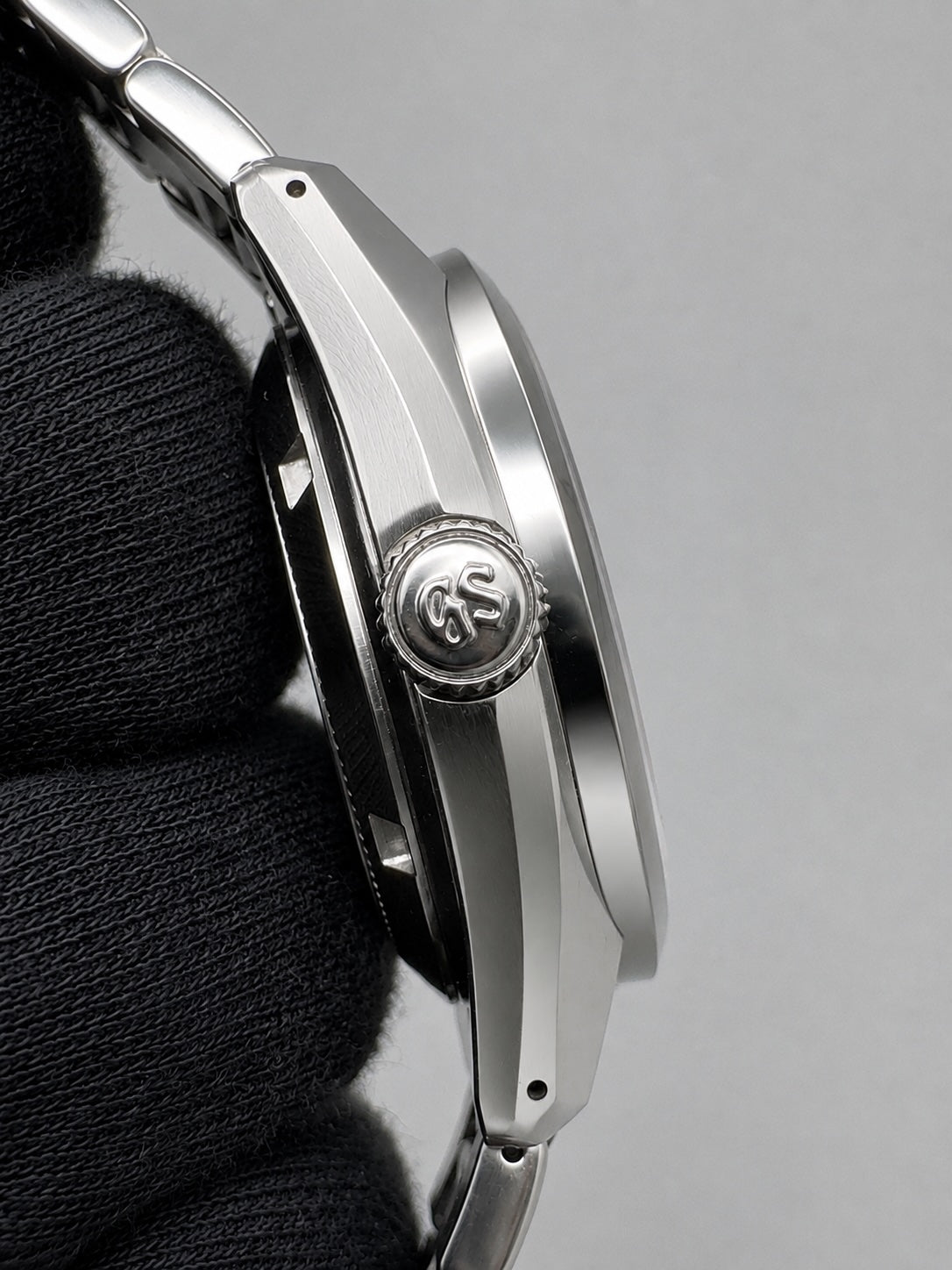 Close-up of a silver stainless steel watch with a matching metal bracelet, held by a black gloved hand against a neutral gray background.