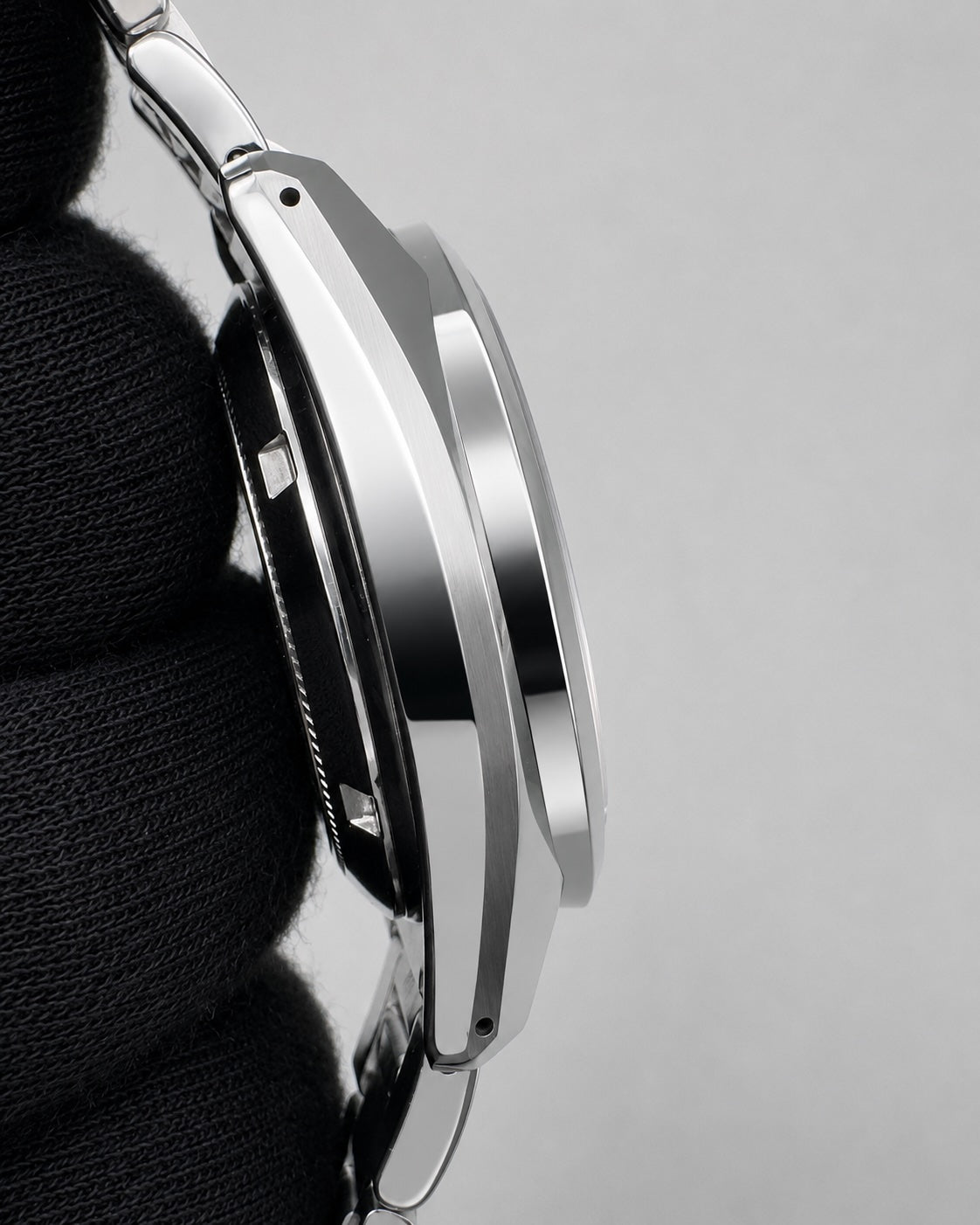 Close-up side profile of a polished silver-toned metal watch case held against a black textured fabric, set against a light gray background.